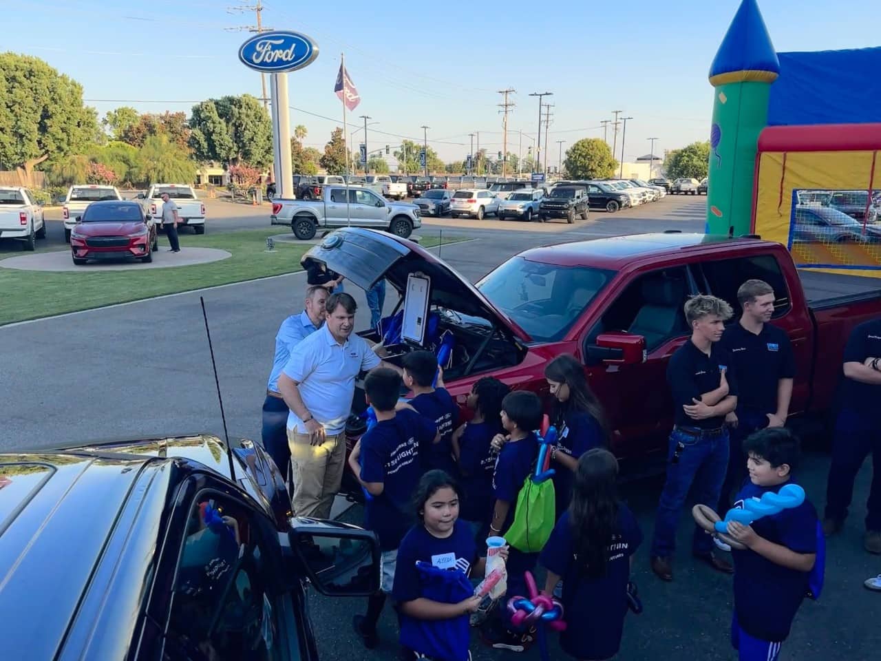 A man hands backpacks and other items out of the frunk of his electric truck to children in the parking lot of a ford dealership.